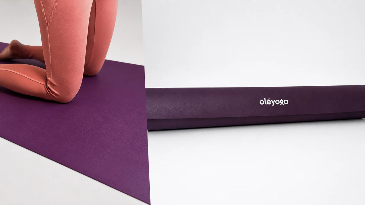 A person kneeling on a purple natural rubber yoga mat, next to the mat rolled on its side