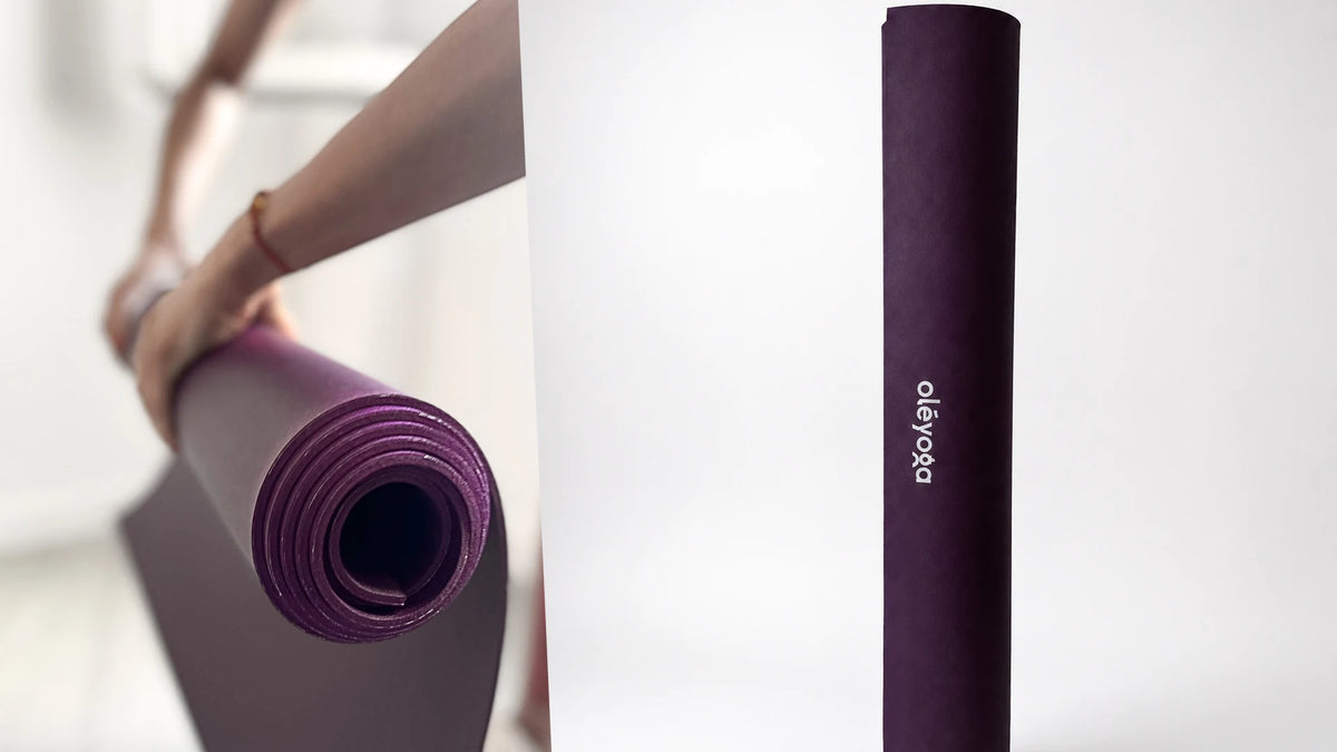 Hands holding a rolled purple natural rubber yoga mat, next to the mat standing upright