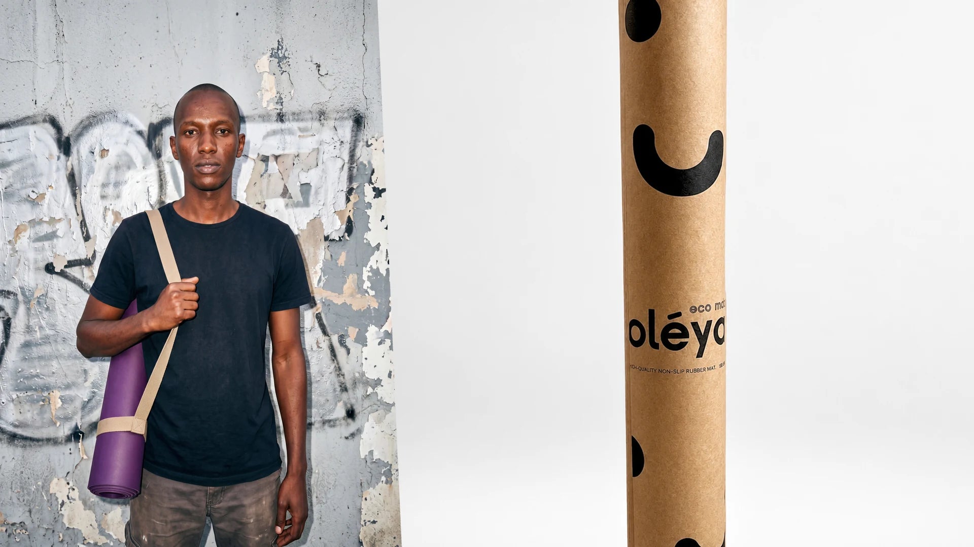Man carrying a rolled purple yoga mat on his shoulder against a graffiti wall, next to the mat in its kraft shipping tube