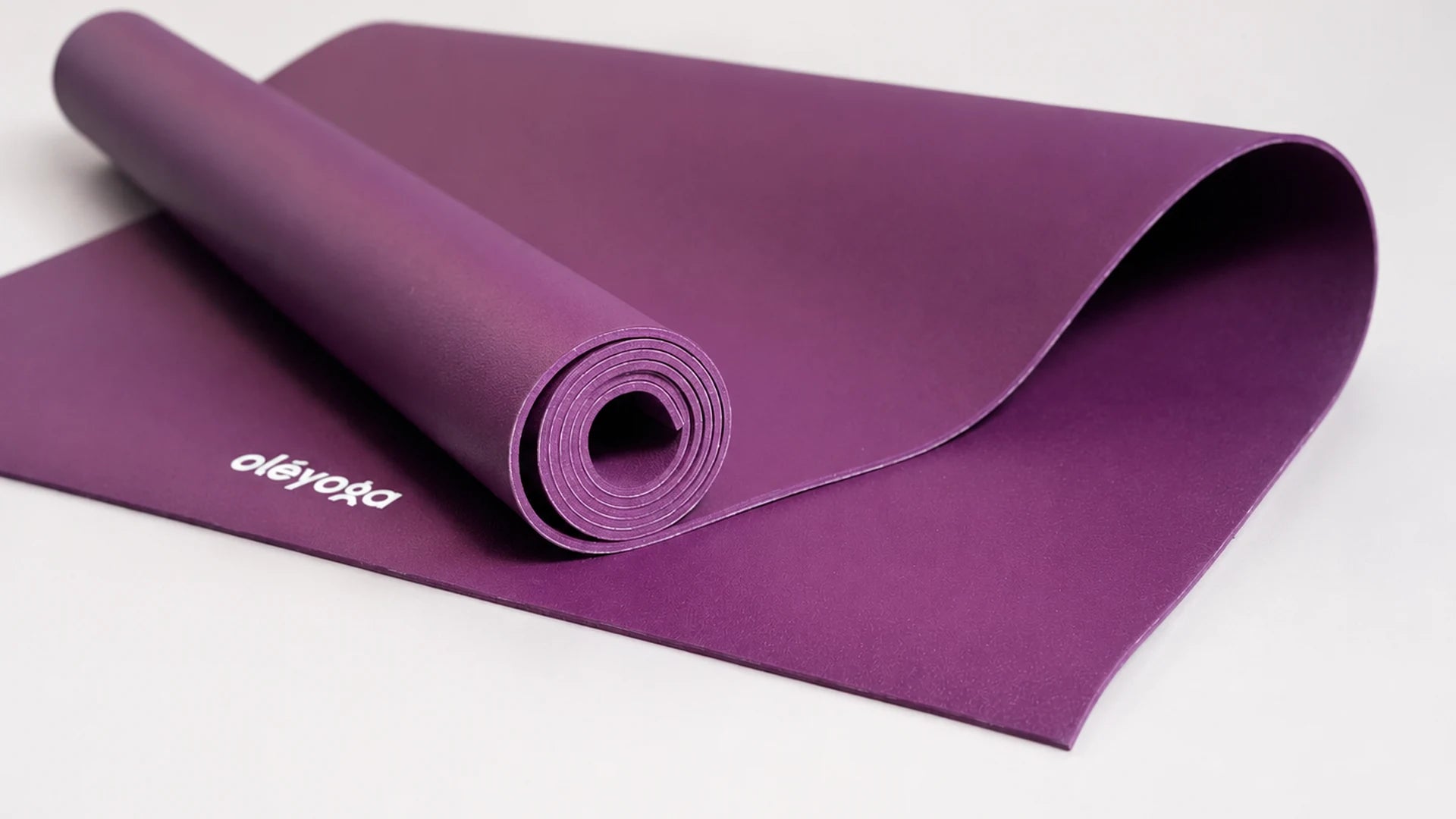 A purple natural rubber yoga mat half unrolled and half rolled up on a white surface, showing the matt surface texture, the edge thickness and the spiral of the roll.