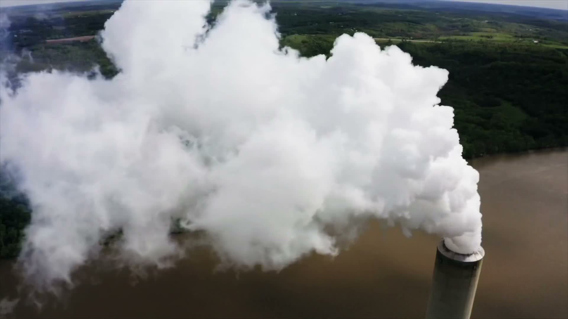 Aerial view of an industrial smokestack emitting a thick plume of smoke over a river and forest